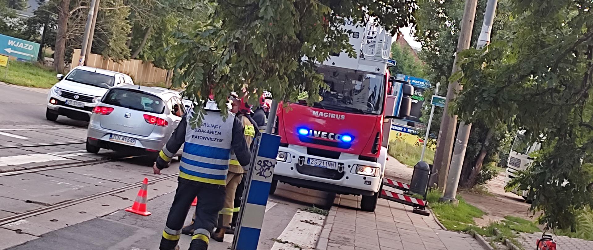 Widać kierującego działaniem ratowniczo gaśniczym odwróconego plecami jest to strażak, w tle widać samochód specjalistyczny z drabina mechaniczna.