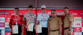 Na podium stoi trzech zawodników biorący udział w wyścigu, po prawej stronie widać dwóch funkcjonariuszy PSP w żółtych ubraniach służbowych. W tle reklamy sponsorów