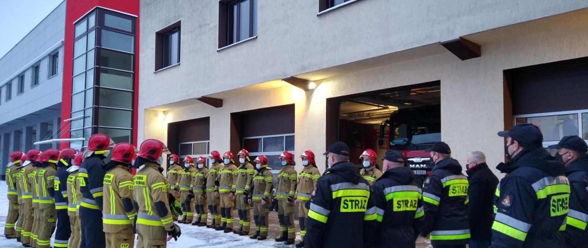 Na zdjęciu widnieją strażacy podczas uroczystej zmiany służby, w której trakcie wręczone zostały grawertony od Ministra Spraw Wewnętrznych i Administracji. Jest dzień, brak słońca, bez opadów, śnieg. 