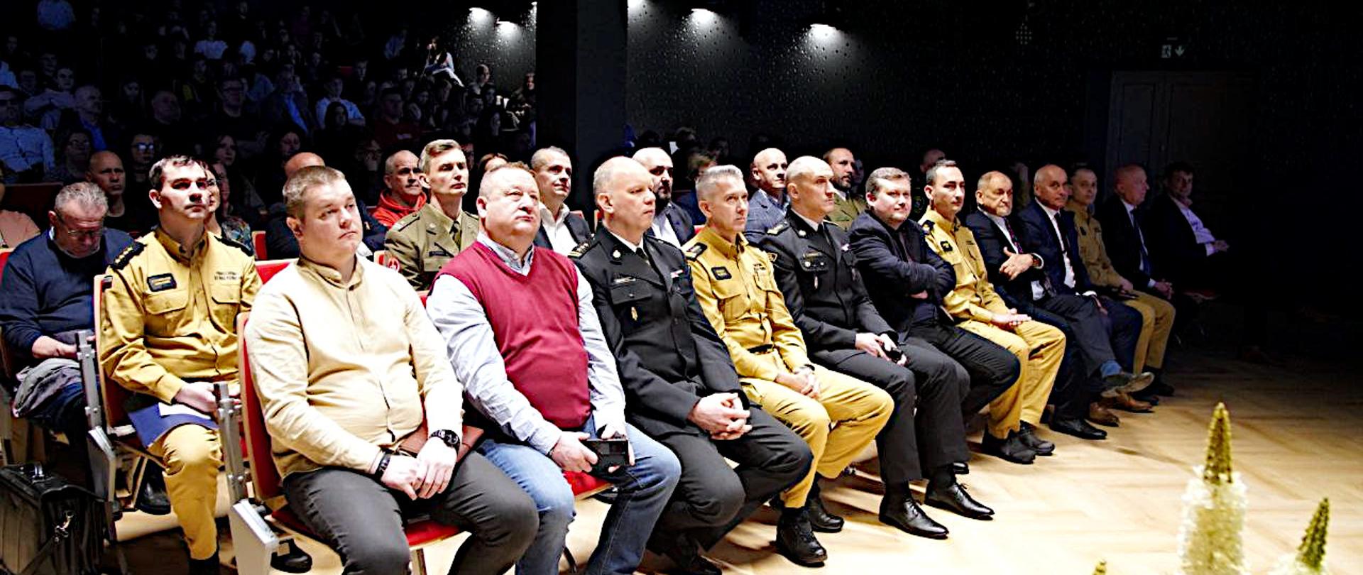 Zdjęcie przedstawia wnętrze dużej, audytoryjnej sali. Publiczność składa się z kilkudziesięciu osób, zarówno mężczyzn, jak i kobiet, w różnym wieku. Siedzą na krzesłach ustawionych w rzędach, wypełniając znaczną część sali. Ich postawa i skupienie sugerują, że uważnie słuchają mówcy. Wśród słuchaczy wyróżnia się grupa mężczyzn w mundurach służbowych strażackich w odcieniach piaskowym wśród nich Zastępca Śląskiego Komendanta Wojewódzkiego PSP st. bryg. Roman Klecha. Oświetlenie sali jest punktowe, skupione na mówcy, Światło z reflektorów tworzy jasny obszar wokół podwyższenia, podczas gdy reszta sali pozostaje w półmroku.