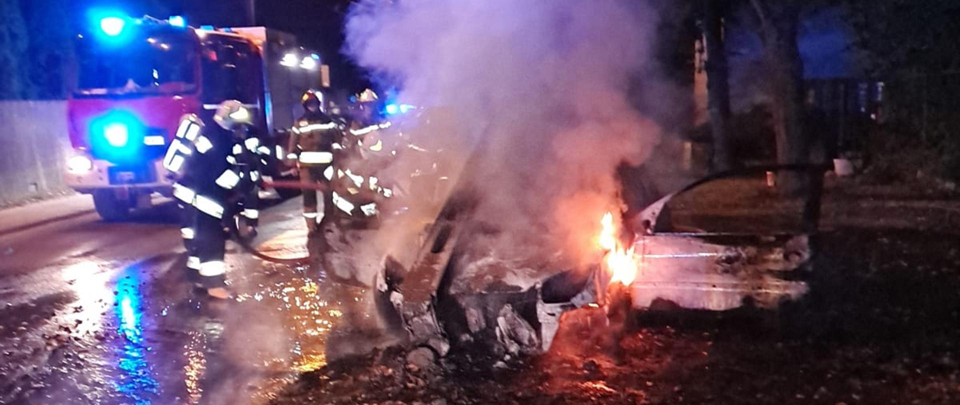 Fotografia przedstawia akcję gaśniczą samochodu osobowego porą nocną. W centralnej części zdjęcia widać palący się samochód, nad którym unosi się siwy dym. Po lewej stronie, na drodze znajdują się strażacy, którzy gaszą pojazd wodą. Za strażakami znajduje się czerwony wóz strażacki, który ma włączone światła alarmowe, koloru niebieskiego. Po prawej stronie rosną drzewa. Trawnik na pierwszym planie zdjęcia jest pełen liści. 