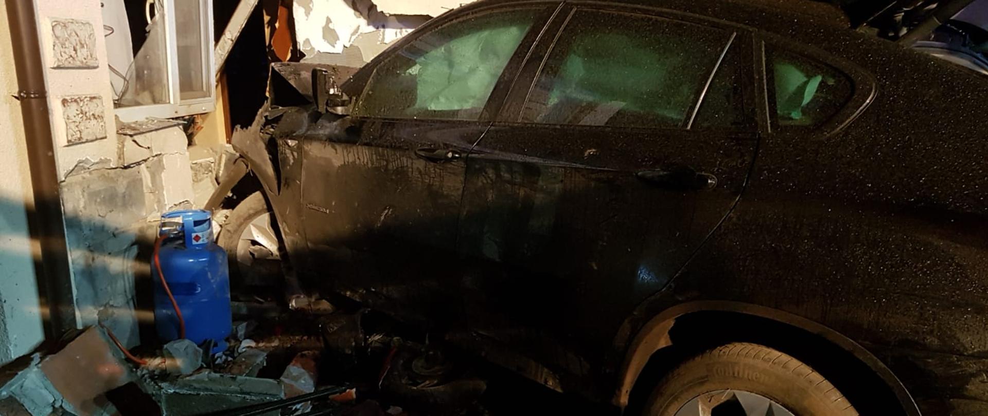 Samochód wjechał w dom uszkadzając jego ścianę, która zawaliła się do środka budynku. Z boku butla z gazem wyniesiona przez strażaków przed dom.