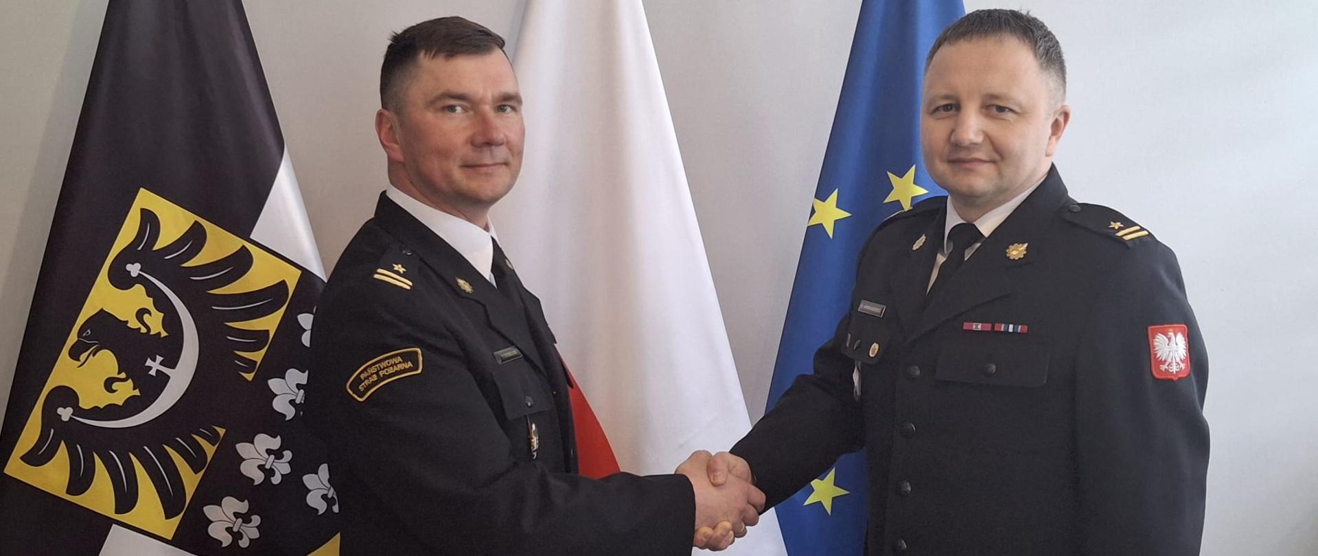 Zdjęcie przedstawia dwóch mężczyzn w ciemnogranatowych mundurach wyjściowych, którzy podają sobie dłonie w uścisku. Stoją na tle trzech flag umieszczonych na stojakach: po lewej stronie znajduje się żółto-czarna flaga Dolnego Śląska z czarnym orłem, w środku flaga Polski, a po prawej stronie niebieska flaga Unii Europejskiej. Nad flagami, na ścianie w tle, wisi niewielki, oprawiony w ramkę obraz lub godło.