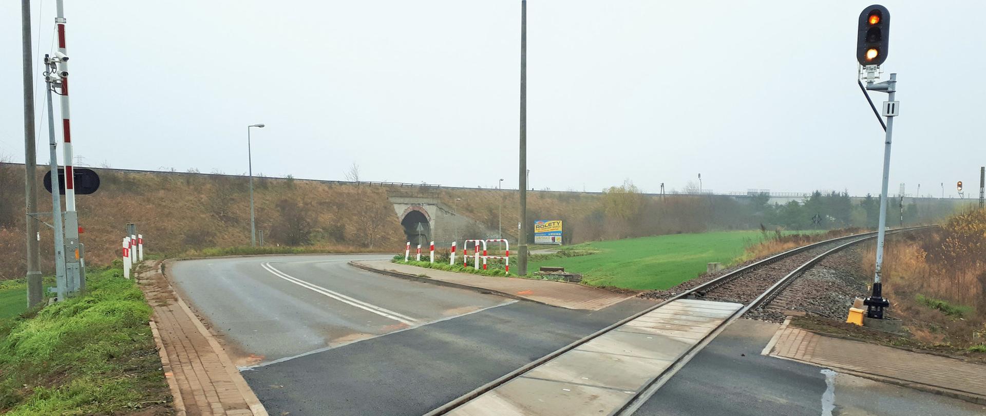 Zdjęcie przedstawia wyremontowany przejazd kolejowy w miejscowości Tczew. Widać po bokach zieloną trawę, a w tle nasyp wraz z drogą.