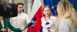 Minister Katarzyna Pełczyńska- Nałęcz stoi w środku przed dziennikarzami, którzy trzymają mikrofony.