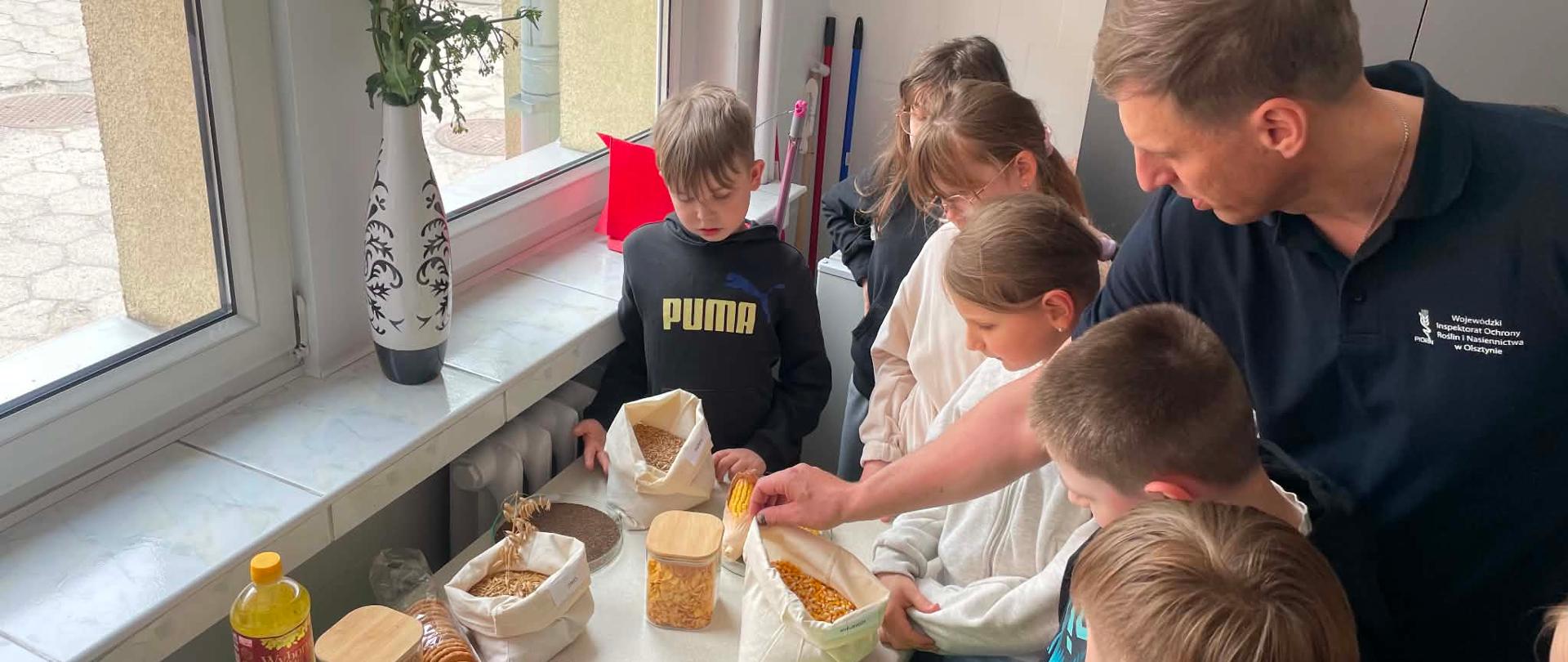 Mężczyzna w koszulce z logo Inspekcji prezentuje uczniom różne rodzaje nasion i produktów roślinnych podczas zajęć edukacyjnych. Dzieci stoją przy stole, na którym znajdują się woreczki z ziarnami oraz produkty roślinne. Spotkanie odbywa się w jasnym pomieszczeniu przy dużym oknie