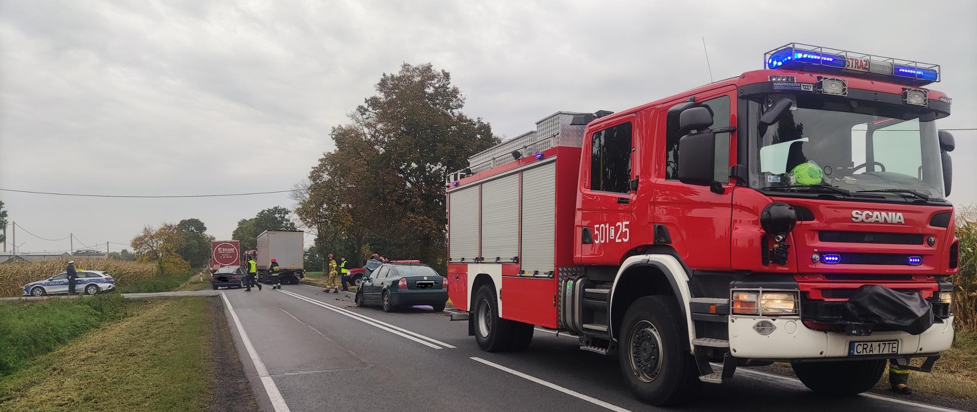 
Zdjęcie przedstawia na pierwszym planie samochód ciężarowy straży pożarnej koloru czerwonego, samochód osobowy oraz ciężarowy z naczepą po kolizji drogowej, radiowóz policyjny, oraz policjantów i strażaków podczas usuwania skutków kolizji drogowej.
