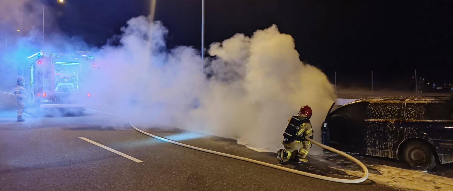 Na zdjęciu po prawej stronie widać fragment spalonego auta pokrytego pianą gaśniczą. Przy nim klęczy ratownik z wężem gaśniczym podający środek gaśniczy na pojazd. W centrum obrazu widzimy kłęby pary wodnej i dymu, a z lewej słabo widoczny stoi samochód pożarniczy oraz ratownik obsługujący autopompę. 