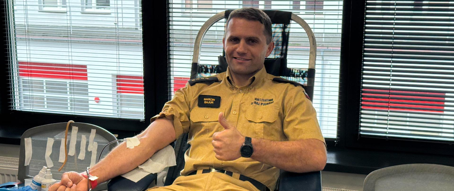 Uśmiechnięty strażak Państwowej Straży Pożarnej, st. kpt. Marcin Bajur, siedzi na leżance medycznej podczas oddawania krwi i pokazuje gest zaciśniętej pięści z uniesionym kciukiem.