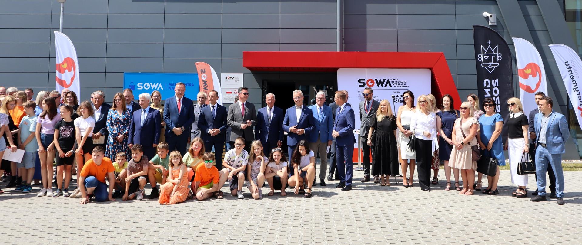 Zdjęcie z odległości, na dworze w ostrym słońcu przed wejściem do szarego budynku z napisem SOWA stoi grupa ludzi, przed nimi na ziemi siedzą dzieci.