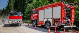 Na zdjęciu na piaskowej drodze leśnej widoczne dwa czerwone samochody pożarnicze. Po prawej stronie drogi widoczny las iglasty.