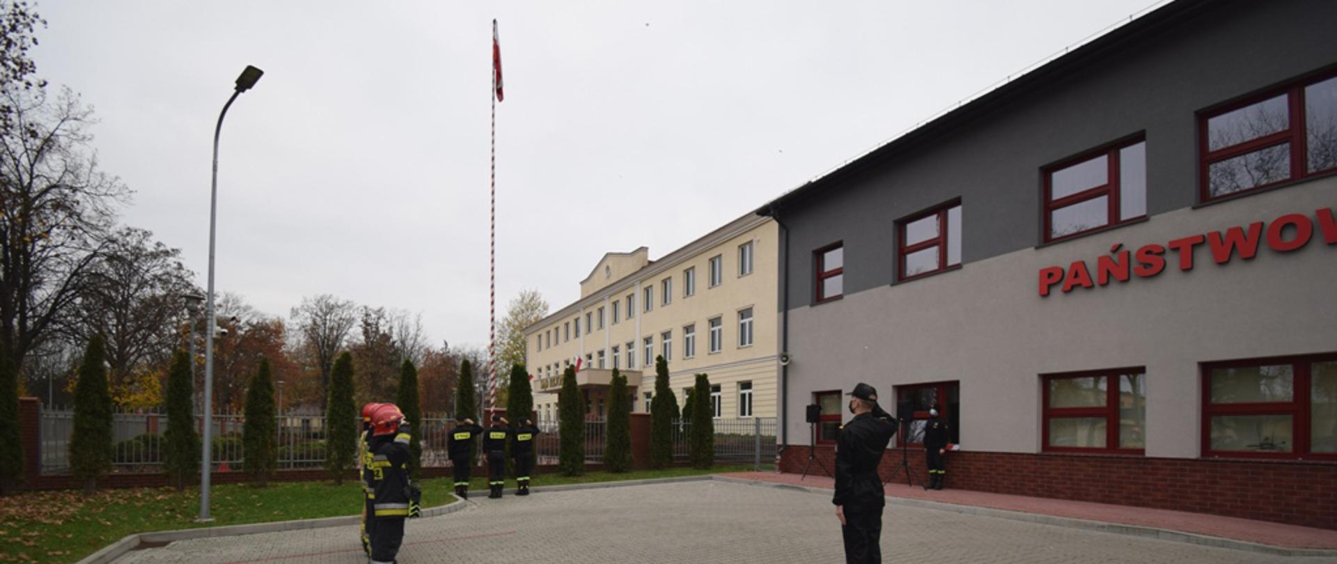podniesienie flagi państwowej, szereg strażaków KP PSP i dowódca, na placu przy komendzie, poczet flagowy, w tle budynek murowany dwukondygnacyjny