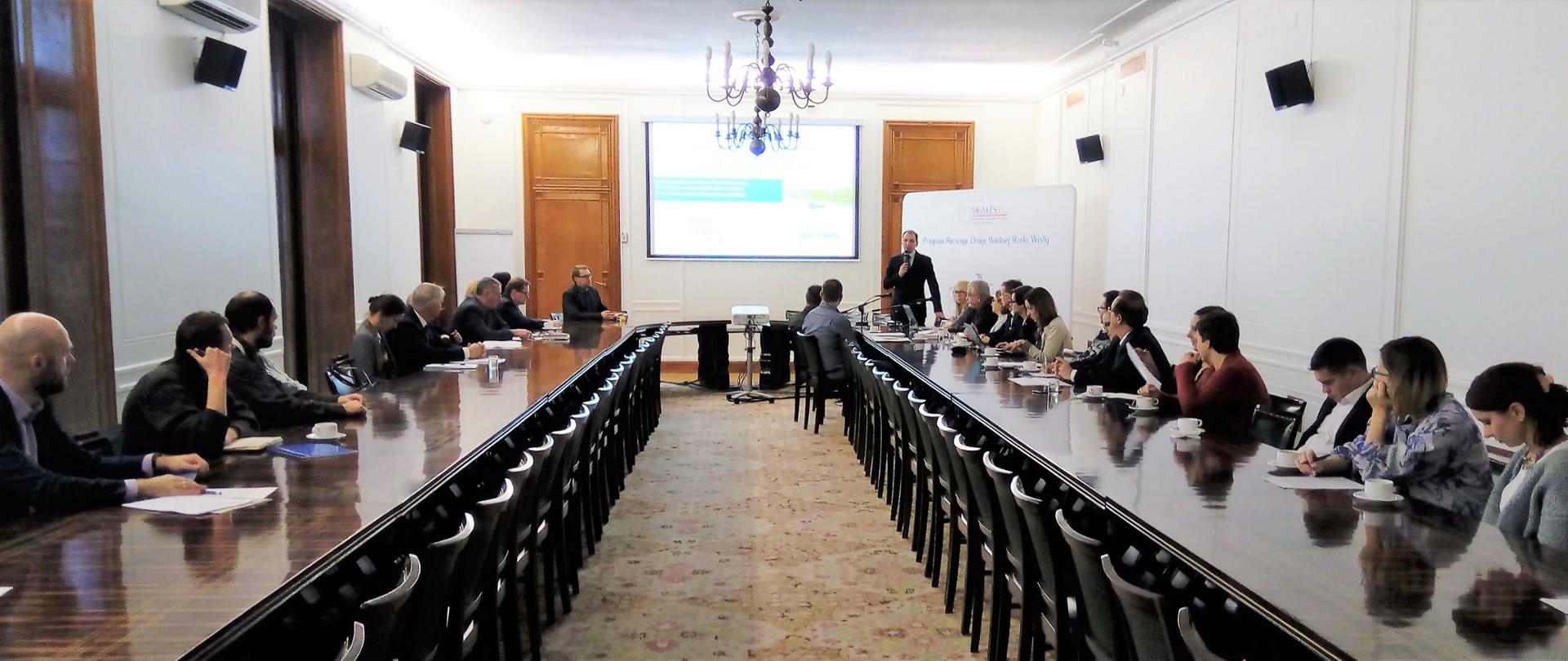 Uczestnicy siedzą przy stole słuchając prelegenta