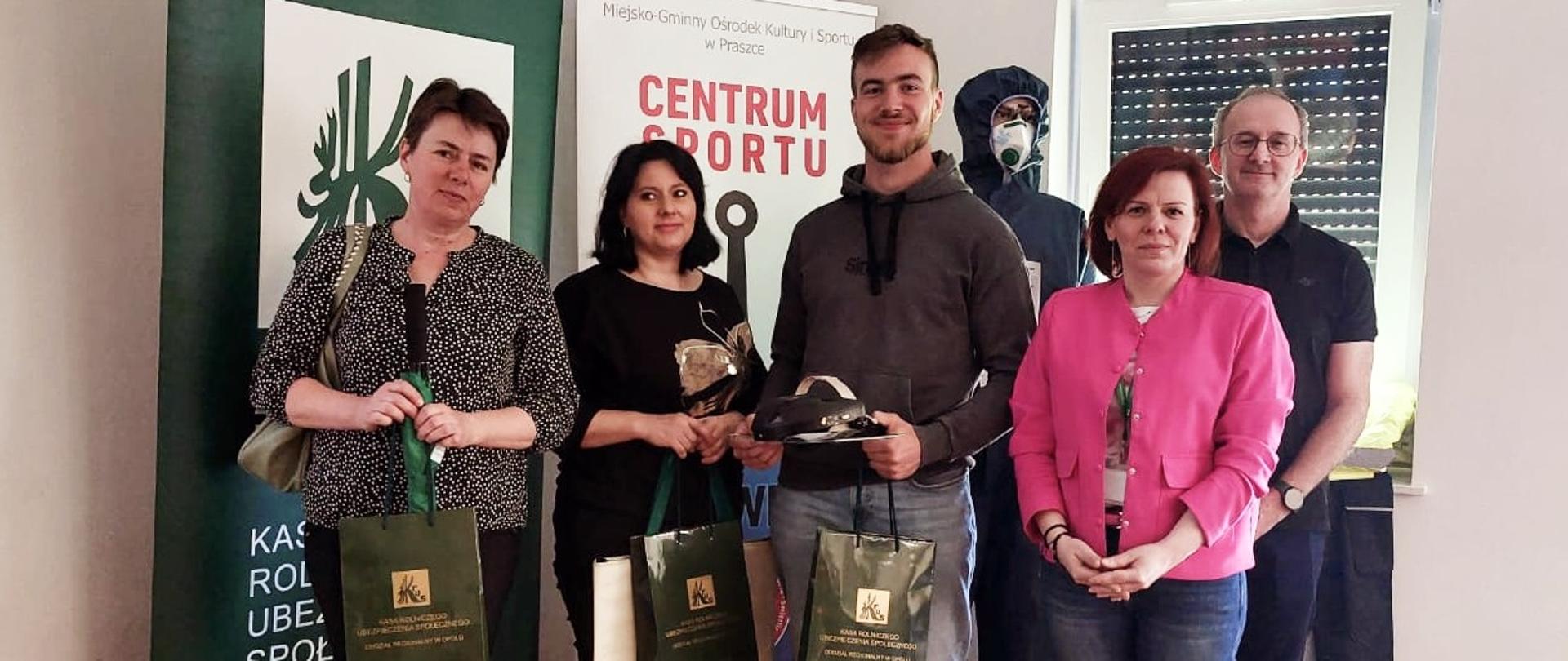 Na tle rollupów z logo KRUS i Centrum Sportu Kotwica stoją dwie kobiety i mężczyzna. Uśmiechają się i pozują do zdjęcia trzymając w rękach zielone torby z logo KRUS z nagrodami. Obok po prawej stoi mężczyzna w okularach i kobieta w różowym żakiecie