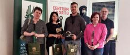 Na tle rollupów z logo KRUS i Centrum Sportu Kotwica stoją dwie kobiety i mężczyzna. Uśmiechają się i pozują do zdjęcia trzymając w rękach zielone torby z logo KRUS z nagrodami. Obok po prawej stoi mężczyzna w okularach i kobieta w różowym żakiecie