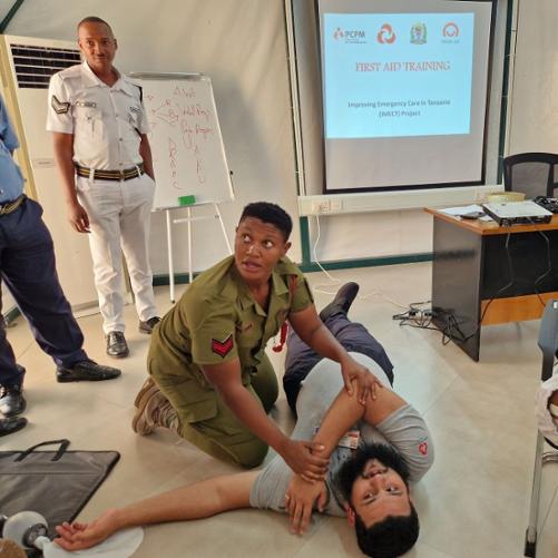 Mężczyzna leży na ziemi, kobieta w mundurze pokazuje na nim zasady pierwszej pomocy