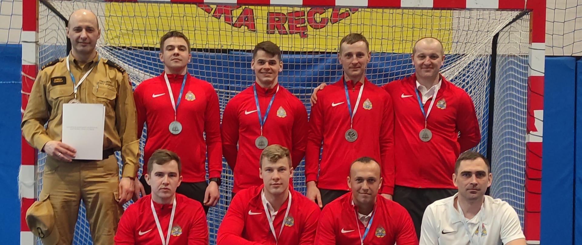 XXI Mistrzostwa Województwa Mazowieckiego Strażaków w Halowej Piłce Nożnej - wspólne zdjęcie po zawodach na sali, w tle bramka.