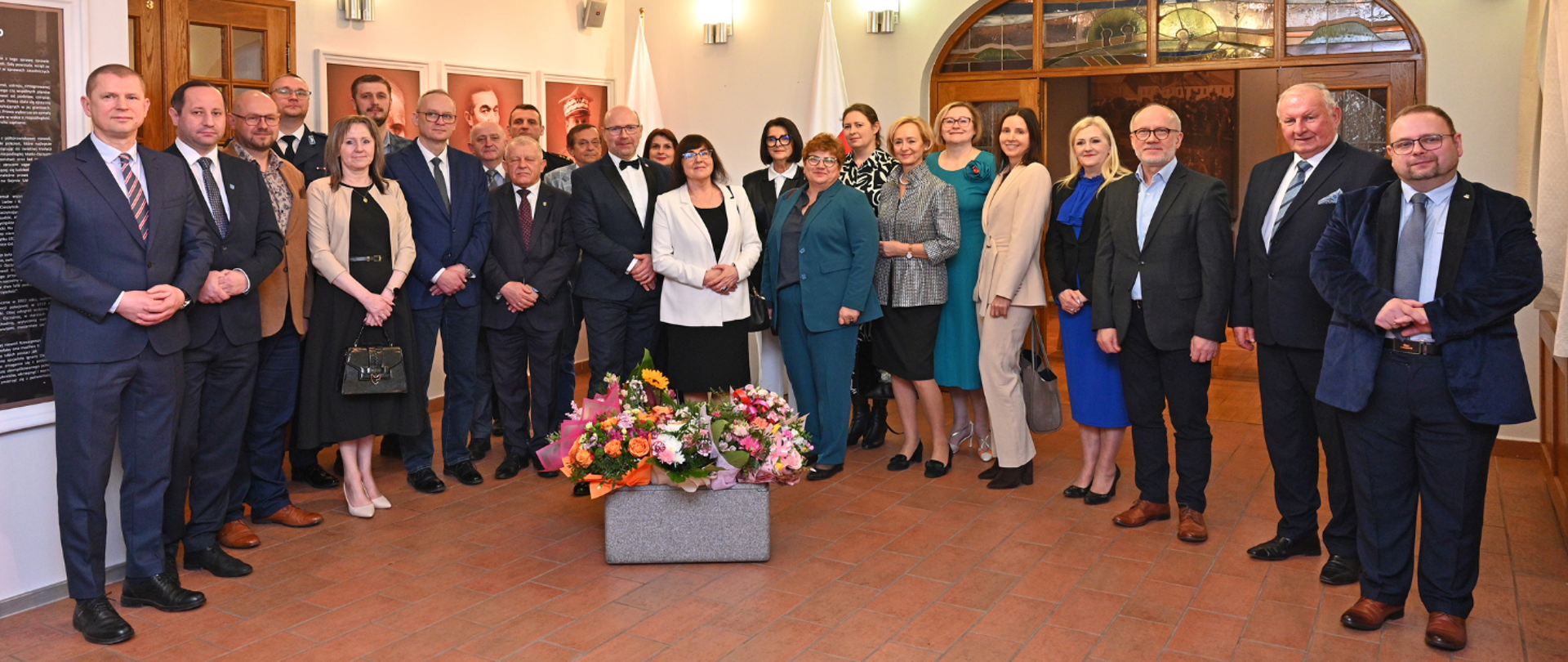 Wspólne, oficjalne zdjęcie grupowe podczas uroczystości, z Ewą Jankowską stojącą centralnie i bukietami kwiatów na pierwszym planie.