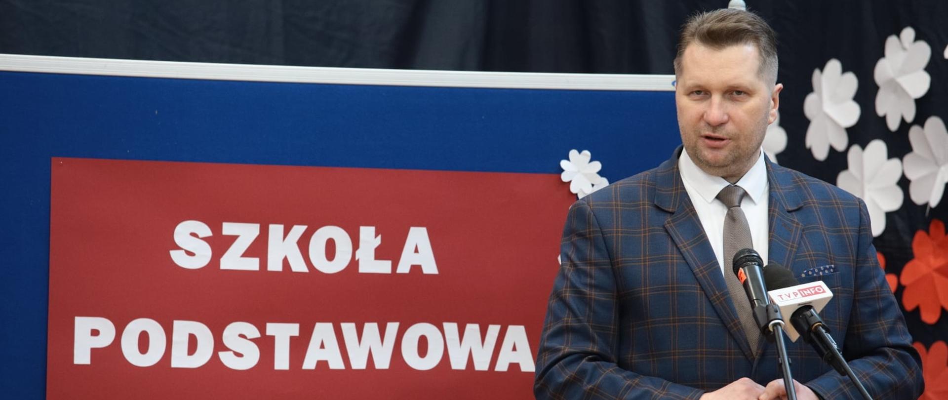 Minister Przemysław Czarnek przemawia podczas uroczystości. Za nim widać napis Szkoła Podstawowa w Snobkowie.