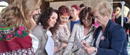 Na pierwszym planie widoczny jest stół z dokumentami, broszurami informacyjnymi, kalkulatorem oraz przyborami biurowymi. Pracownica Oddziału Promocji Zdrowia i Komunikacji Społecznej zapoznaje pozostałych uczestników z treścią dokumentu, który trzyma w dłoni. Wokół stanowiska zgromadzonych jest kilka osób, pochylonych nad materiałami i uważnie śledzących przekazywane informacje, co wskazuje na konsultacyjny i edukacyjny charakter rozmowy. 