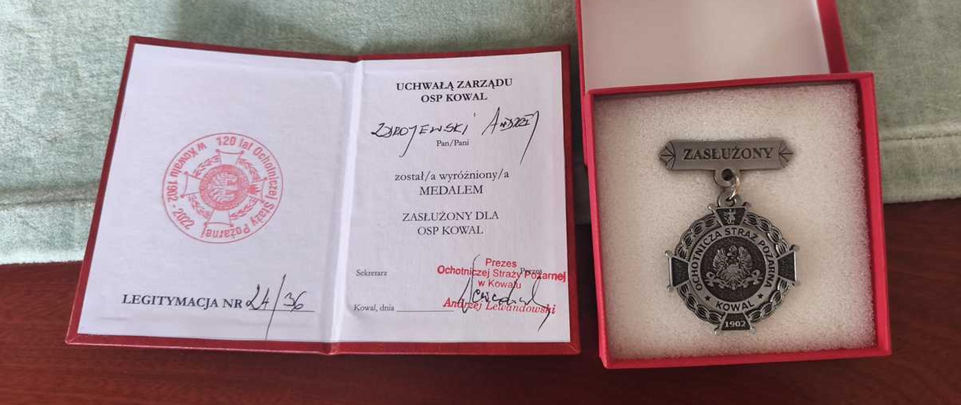 "Zdjęcie przedstawia otwartą legitymację oraz medal w etui, leżące na ciemnoczerwonym stole na tle jasnego, miękkiego materiału.
• Po lewej stronie znajduje się otwarta legitymacja papierowa z białymi stronami i czerwoną ramką.
• Na lewej stronie widoczny jest okrągły, czerwony stempel okolicznościowy z napisem „120 lat Ochotniczej Straży Pożarnej w Kowalu 1902-2022”. Na dole strony widnieje odręczny napis „LEGITYMACJA NR 24/36”.
• Na prawej stronie dokumentu znajduje się nagłówek „UCHWAŁĄ ZARZĄDU OSP KOWAL”. Poniżej wpisane jest odręcznie nazwisko i imię: „Zdrojewski Andrzej”. Tekst drukowany głosi: „został/a wyróżniony/a MEDALEM ZASŁUŻONY DLA OSP KOWAL”. Na dole strony widnieją podpisy oraz czerwona, podłużna pieczątka: „Prezes Ochotniczej Straży Pożarnej w Kowalu Andrzej Lewandowski”.
• Po prawej stronie widoczne jest otwarte, kwadratowe etui w kolorze czerwonym z jasnym, gąbkowym wypełnieniem w środku. Spoczywa w nim srebrno-czarny, metalowy medal.
• Górna część medalu to podłużna baretka z napisem „ZASŁUŻONY”.
• Od baretki, na małym łańcuszku, wisi okrągły medalion. W jego centralnej części znajduje się znak Ochotniczej Straży Pożarnej, a wokół niego biegnie napis „OCHOTNICZA STRAŻ POŻARNA • KOWAL • 1902”."