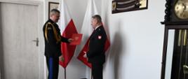 Pomorski komendant wojewódzki Państwowej Straży Pożarnej wręcza czerwoną teczkę funkcjonariuszowi za strażakami znajdują się flagi Polski.