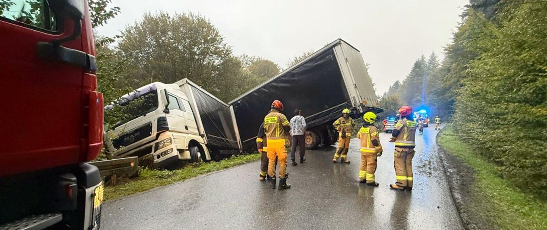 Zdjęcie przedstawia wypadek drogowy w miejscowości Kwaszenina. Ciężarówka (ciągnik siodłowy z naczepą) zjechała z drogi i znajduje się częściowo w rowie. Naczepa jest przechylona. Na miejscu znajduje się kilku strażaków w pełnym umundurowaniu, którzy zabezpieczają teren i prawdopodobnie oceniają sytuację. W tle widać radiowozy i inne pojazdy służb ratunkowych z włączonymi światłami sygnalizacyjnymi. Droga jest mokra, co może wskazywać na deszczową pogodę i śliską nawierzchnię – możliwy czynnik przyczyniający się do wypadku. Otoczenie to zalesiony teren, droga jest wąska i kręta.