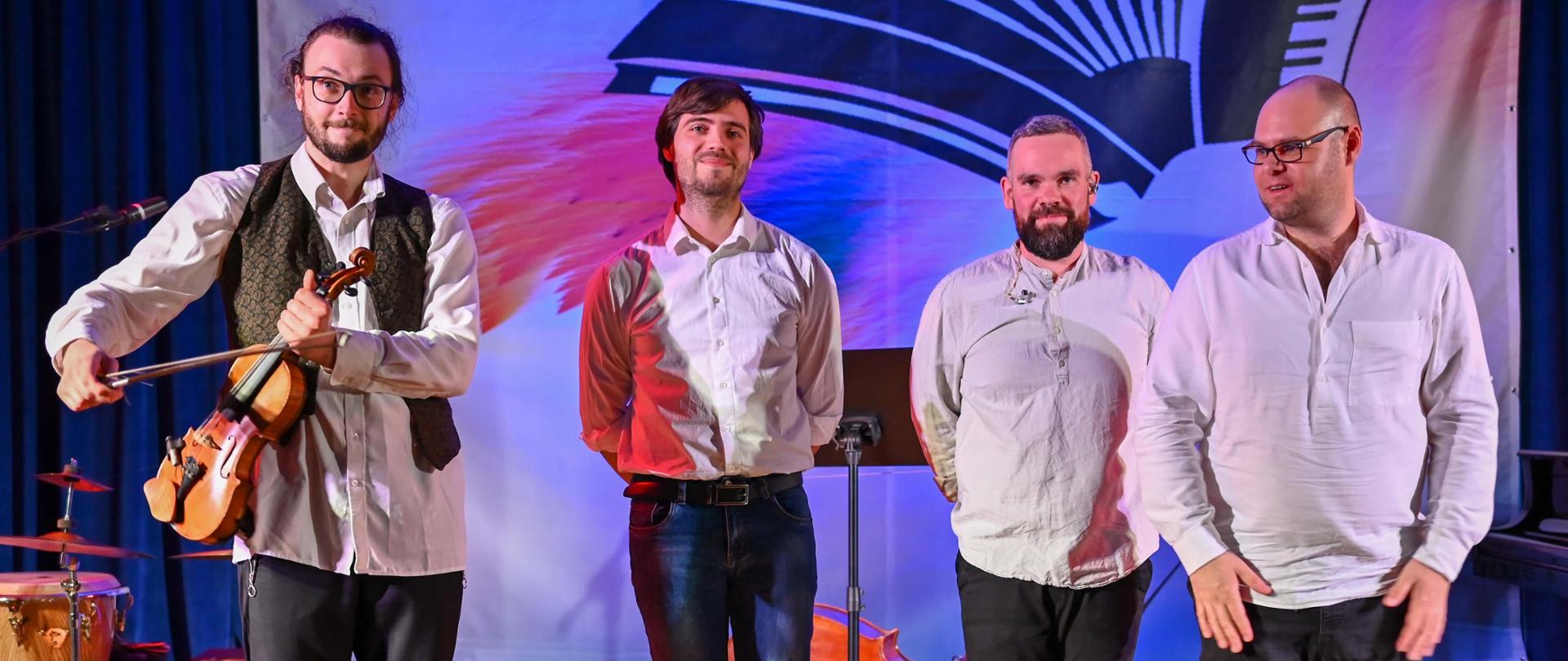 grupa czterech muzyków w białych koszulach stojąca na scenie, za nimi instrumenty, baner konkursowy i niebieska kotara