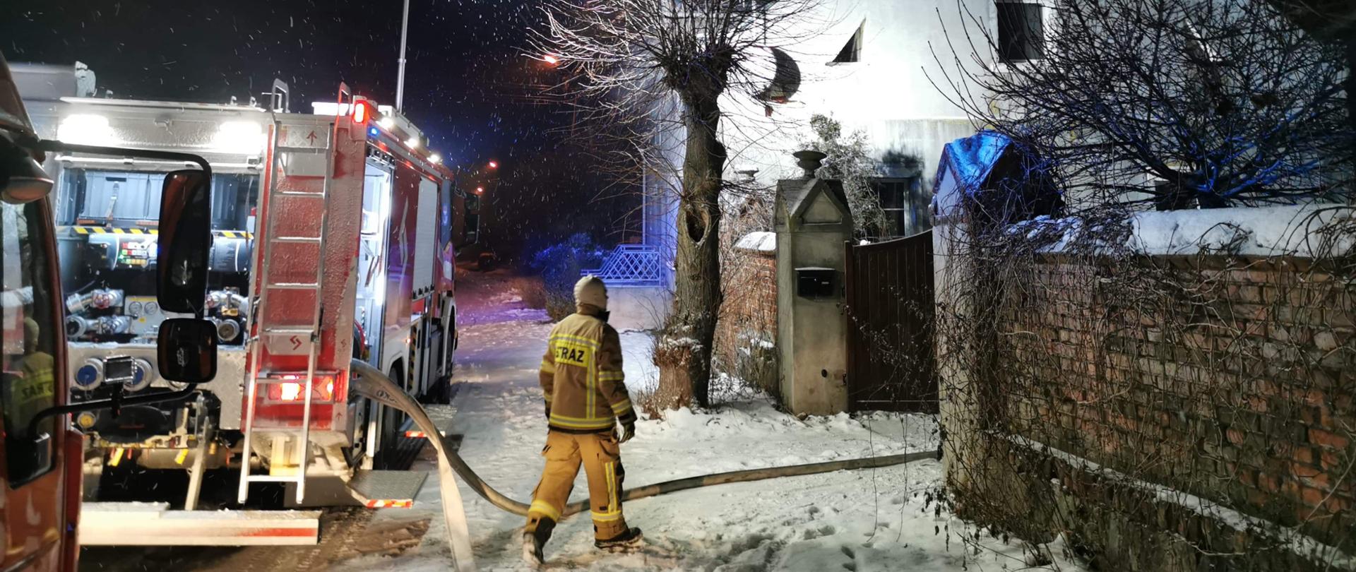 Pora nocna. Śnieg na drodze. Wozy strażackie stoją przed jasnym domem. Strażak w jasnym ubraniu idzie. Dom oświetlony światłem. Obok szary mur z cegieł.
