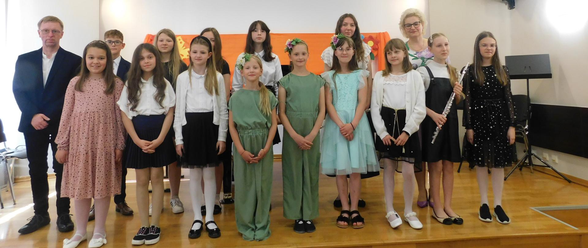 Na zdjęciu grupa uczniów- chłopcy i dziewczęta. Wszyscy ubrani odświętnie stoją na drewnianej scenie. w tle za uczniami pomarańczowa płachta zawieszona na ścianie. Wśród uczniów dwóch nauczycieli mężczyzna- blondyn ubrany w ciemnym garniturze i białej koszuli oraz kobieta- blondynka w okularach. 