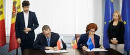Kobieta i mężczyzna w oficjalnych strojach podpisujący dokumenty przy flagach Polski, Mołdawii, Unii Europejskiej i UNDP.