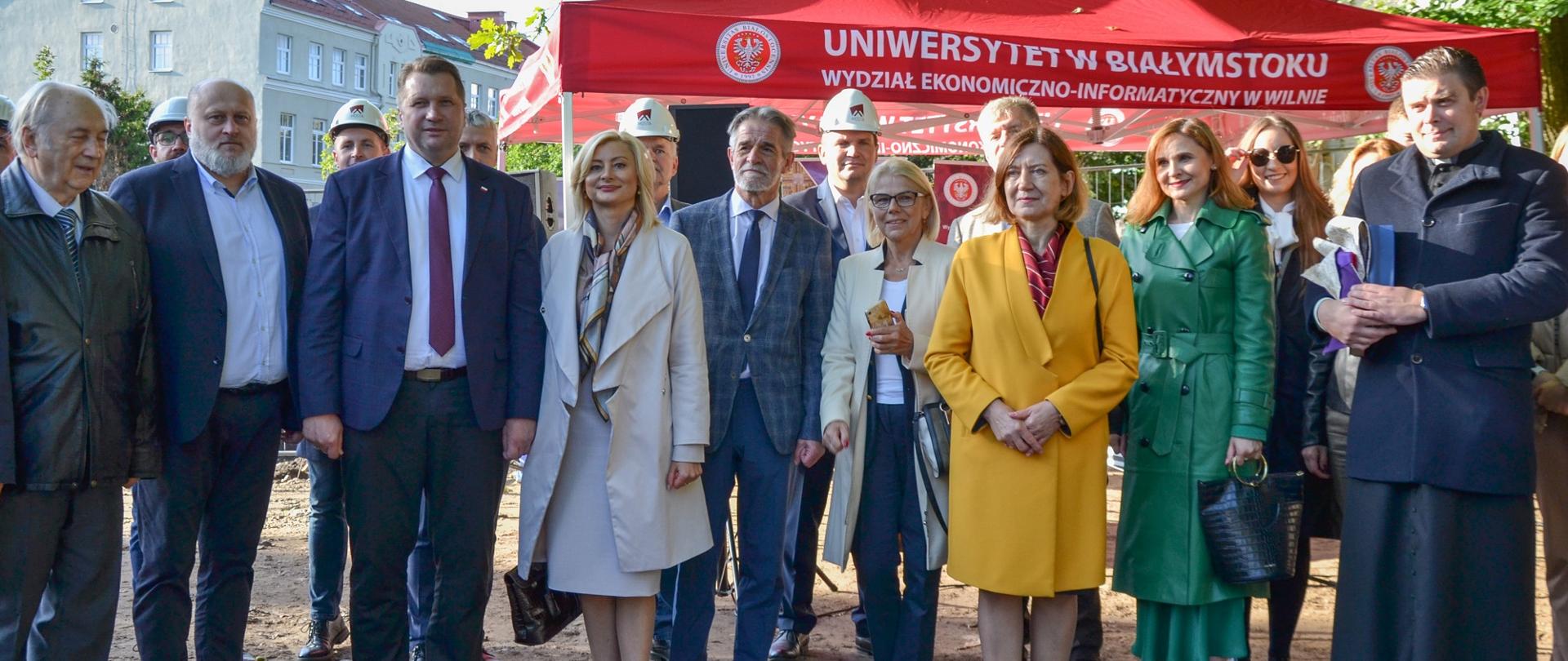 Zdjęcie zbiorowe - minister Czarnek w otoczeniu grupy ludzi stoi przed czerwonym namiotem, w tle miejskie zabudowania.