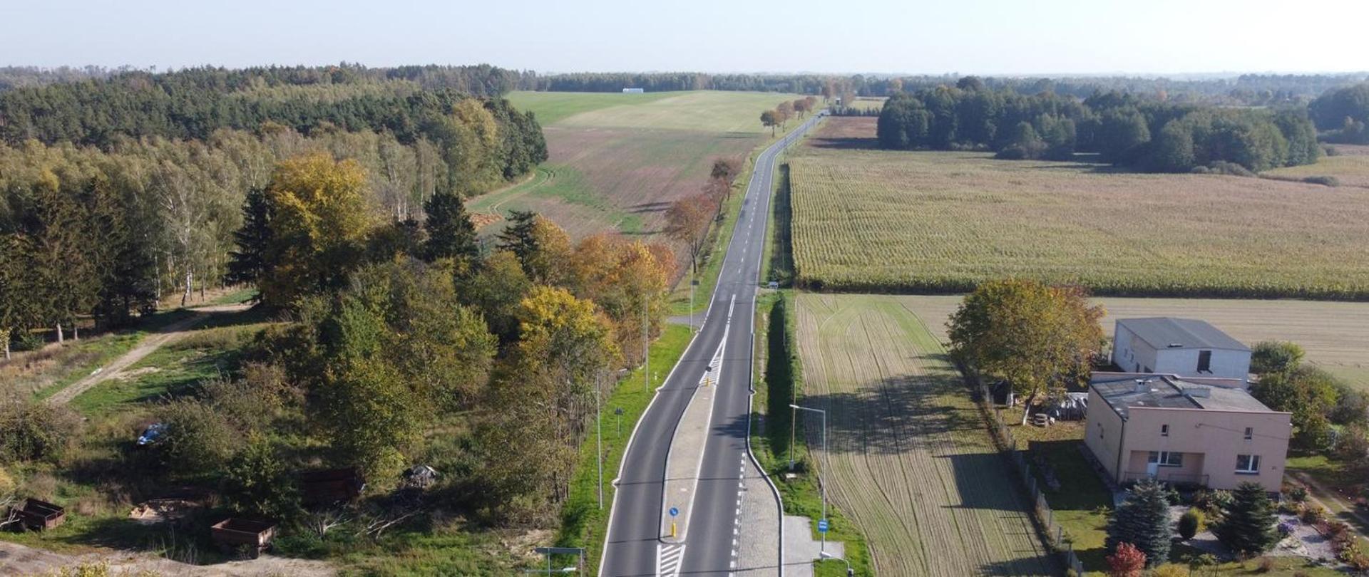 Widok z lotu ptaka na nowo wybudowany odcinek drogi krajowej przebiegający przez tereny rolnicze, z widocznym oznakowaniem poziomym, poboczem oraz zabudową i zadrzewieniem po bokach.