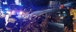 Uszkodzony samochód osobowy pod zderzeniu z ciężarówką. Pojazd ma uszkodzony przód oraz bok. Wokół niego stoją strażacy prowadzący działania w ubraniach bojowych. W tle samochód pożarniczy z załączonymi sygnałami.