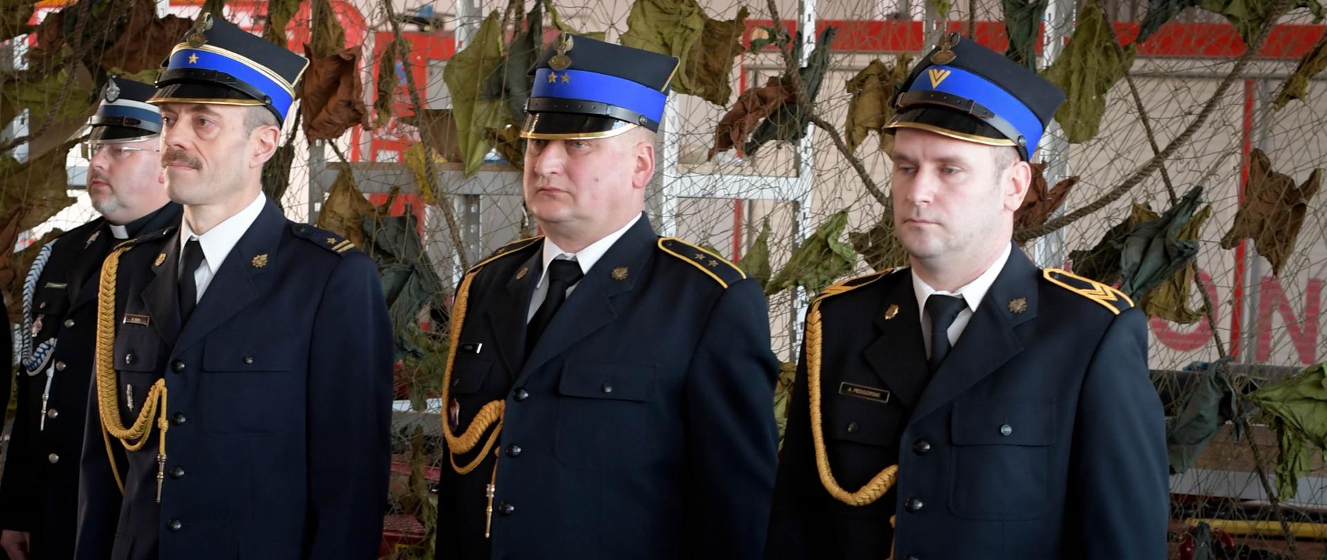 Zdjęcie przedstawia sylwetki trzech strażaków odchodzących na zaopatrzenie emerytalne, ubranych w mundury wyjściowe w postawie zasadniczej. Strażacy stoją na tle siatki maskującej w garażu Jednostki Ratowniczo-Gaśniczej Nr 3 w Rzeszowie.