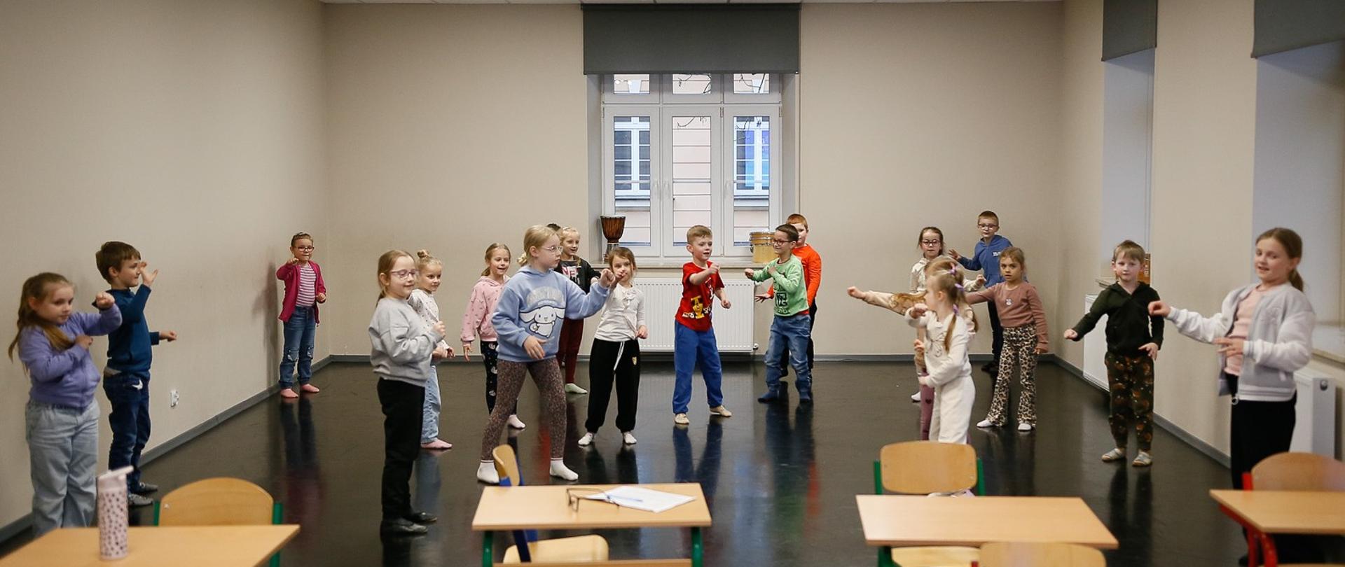 Grupa dzieci w wieku przedszkolnym i wczesnoszkolnym uczestniczy w zajęciach rytmiki w jasnej sali. Dzieci stoją w rozproszeniu, wykonując dynamiczne gesty rękami w rytm muzyki.