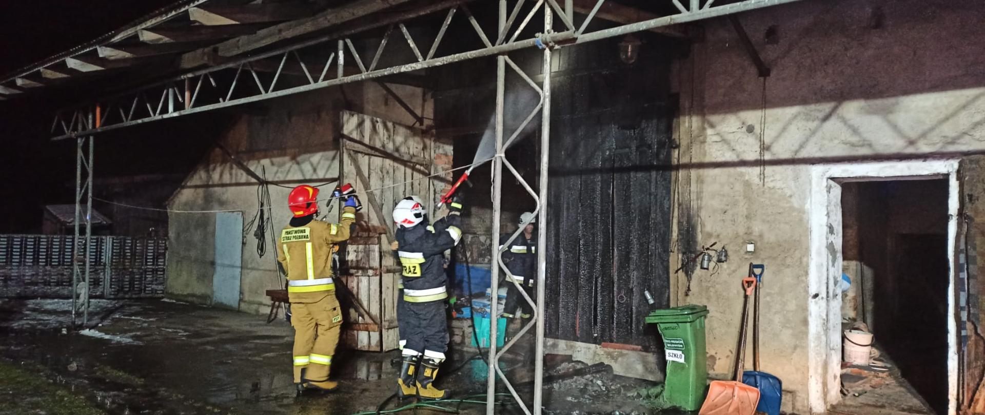 Na zdjęciu strażacy gaszą wodą palące się wrota do stodoły. Zdjęcie wykonane porą nocną.