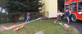 Z prawej samochód pożarniczy czerwony ciężarowy, z samochodu rozwinięta jest linia szybkiego natarcia, z której przewracane są przez chłopca pachołki prądem wody. 