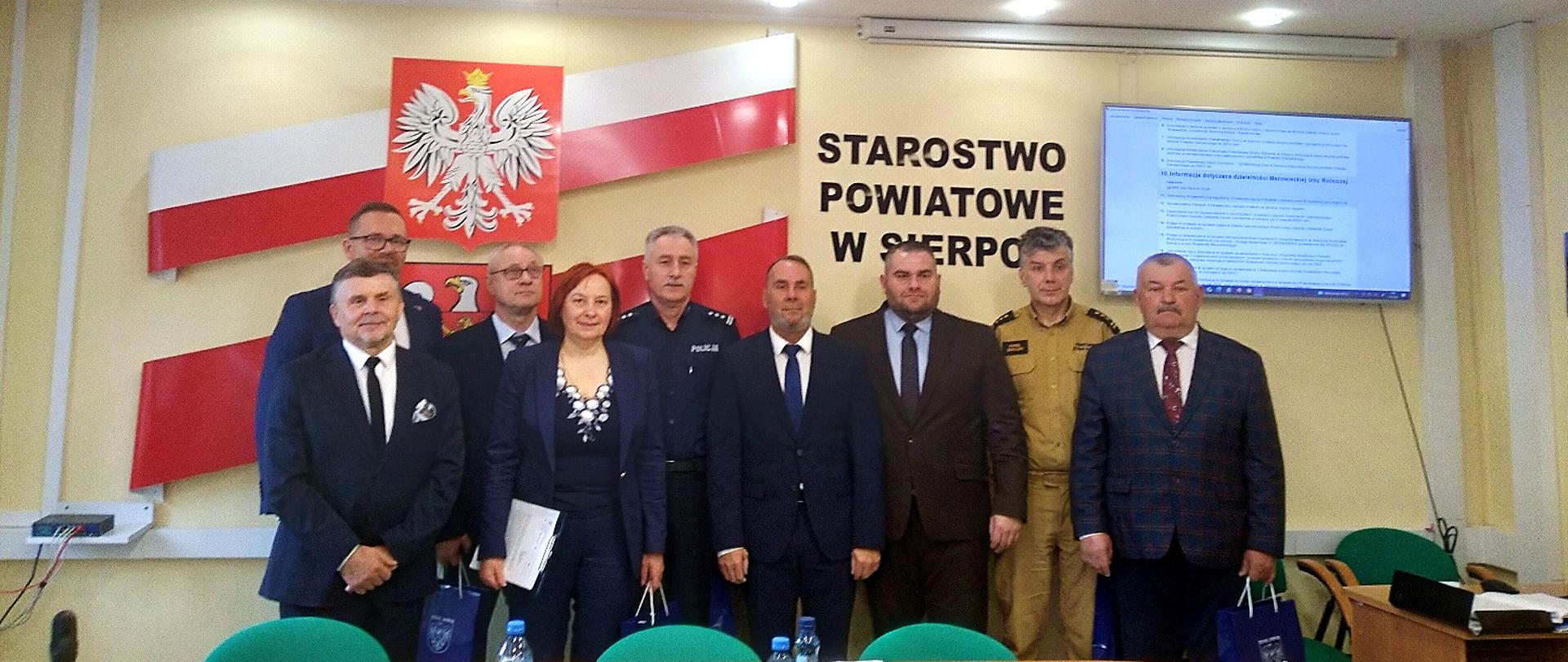 Na zdjęciu widać uczestników Sesji powiatu Sierpeckiego, pozujących do wspólnej fotografii w sali konferencyjnej Starostwa Powiatowego w Sierpcu. W tle widać godło Polski, herb powiatu sierpeckiego oraz napis "STAROSTWO POWIATOWE W SIERPCU". Obecni są przedstawiciele różnych instytucji, w tym Starosta powiatu oraz Pani Agnieszka Wąs, przedstawicielka Wojewódzkiego Inspektoratu Ochrony Roślin i Nasiennictwa w Warszawie, Oddziału w Płocku.