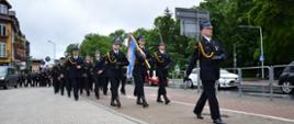Fotografia przedstawia strażaków Państwowej i Ochotniczej Straży Pożarnej maszerujących w mundurach wyjściowych ulicą Mińska podczas Powiatowego Dnia Strażaka. Pierwszy idzie dowódca pododdziału, za nim poczet sztandarowy, dalej pododdział strażaków w czteroszeregu