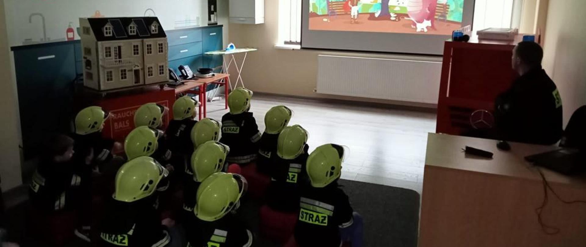 Na zdjęciu grupa przedszkolaków, która odwiedziła wolsztyńską jednostkę. Dzieci siedzą na dywanie i oglądają film edukacyjny.