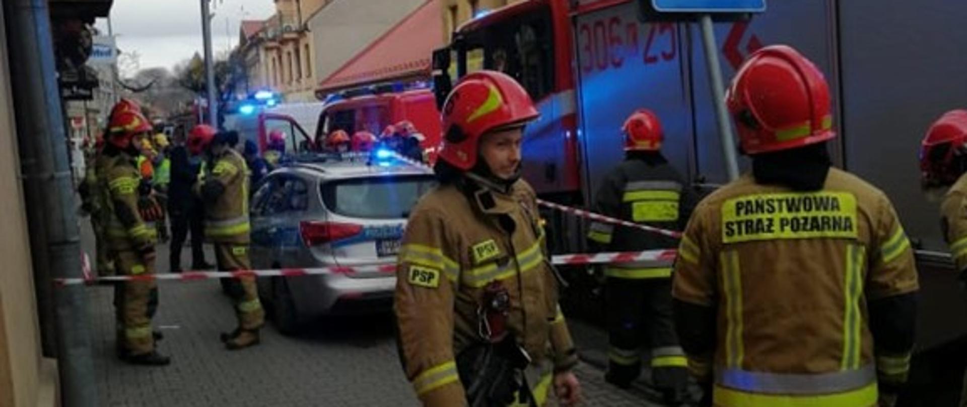 Na zdjęciu po prawej widać samochody pożarnicze wraz z policją i pogotowiem gazowym. Obok samochodów widzimy przechodzących i stojących strażaków pracujących na miejscu zdarzenia. Wzdłuż chodnika przy samochodach pożarniczych oraz w poprzek chodnika jest rozwinięta taśma ostrzegawcza a także rozstawione stożki ostrzegawcze. Przed nimi stoją strażacy dodatkowo zabezpieczający miejsce akcji. 