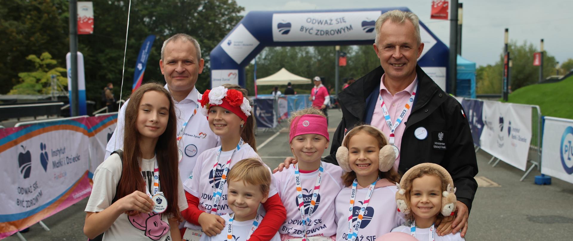 Na asfaltowym placu stoi grupa sześciorga dzieci, za nimi stoi dwóch mężczyzn, w tle zielone drzewa.