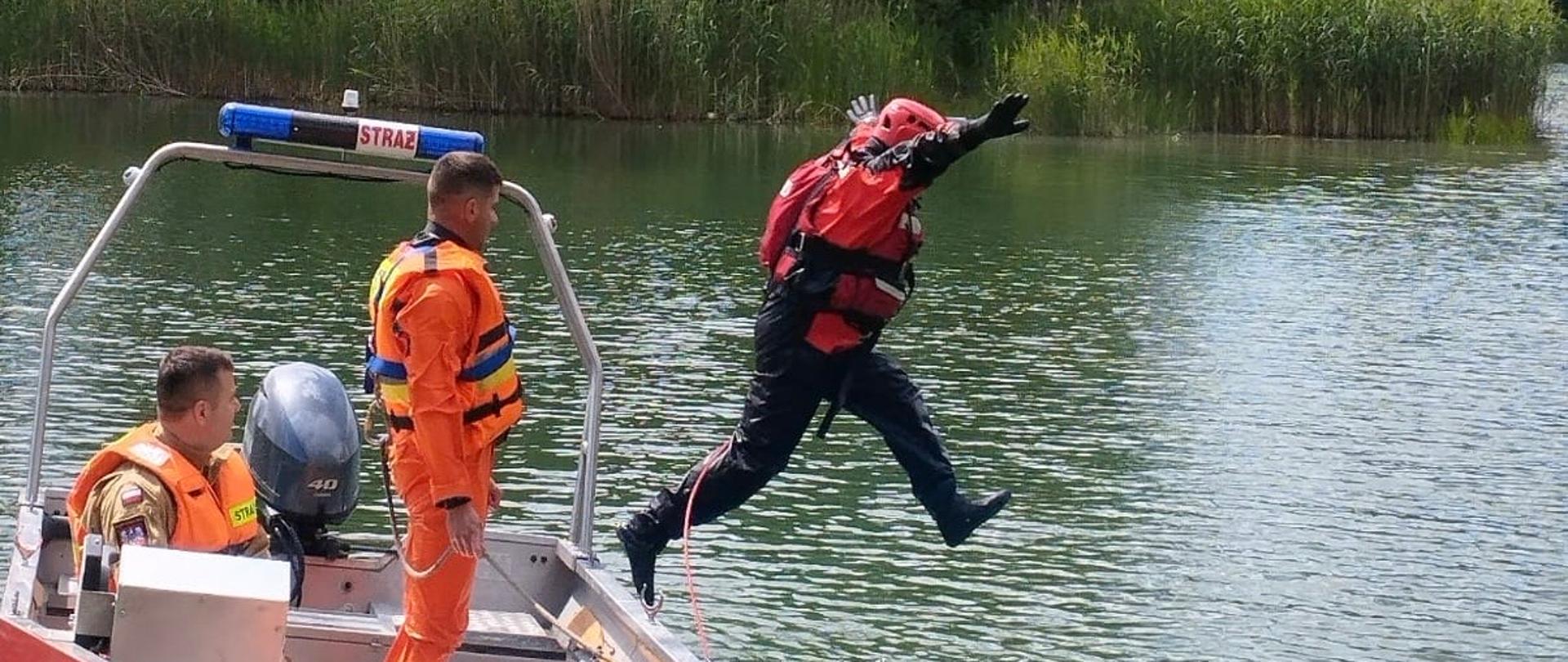 Strażak w specjalistycznym ubraniu do ratownictwa wodnego wykonuje skok z łodzi ratowniczej do wody podczas ćwiczeń. Na pokładzie znajdują się dwaj strażacy zabezpieczający działania.