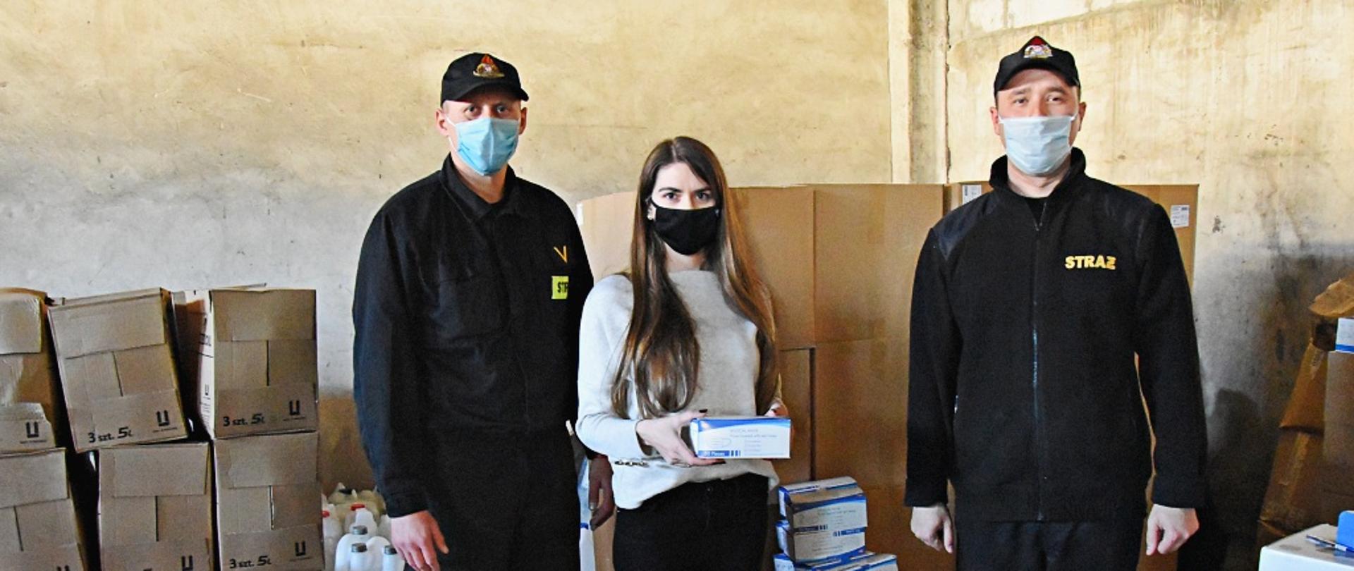 W garażu Jednostki Ratowniczo Gaśniczej w Kutnie stoi dwóch strażaków w czarnych mundurach koszarowych i maseczkach na twarzy. Pomiędzy nimi przedstawicielka oświaty w białej bluzce i czarnych spodniach, trzyma opakowanie z maseczkami ochronnymi. Na twarzy również ma maseczkę ochronną. W tle za nimi stoją opakowania kartonowe koloru szarego ze środkami dezynfekcyjnymi oraz maseczkami.