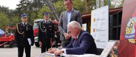 Goście wpisują się do księgi pamiątkowej Komendy Powiatowej PSP w Łowiczu, w tle stoi komendant powiatowy psp w Łowiczu oraz prezesa zarządu powiatowego osp, za nimi drabina mechaniczna oraz zabytkowy wóz straży pożarnej na którym stoi duża statuetka św. Floriana