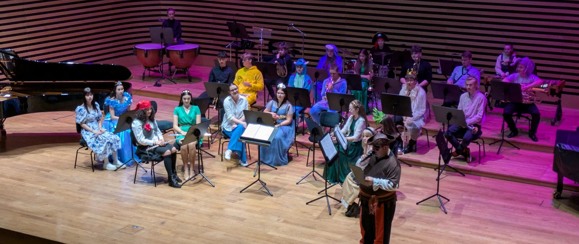 Mężczyzna w stroju pirata zapowiada wykonawców, za nim siedzi orkiestra dęta i kobieta.