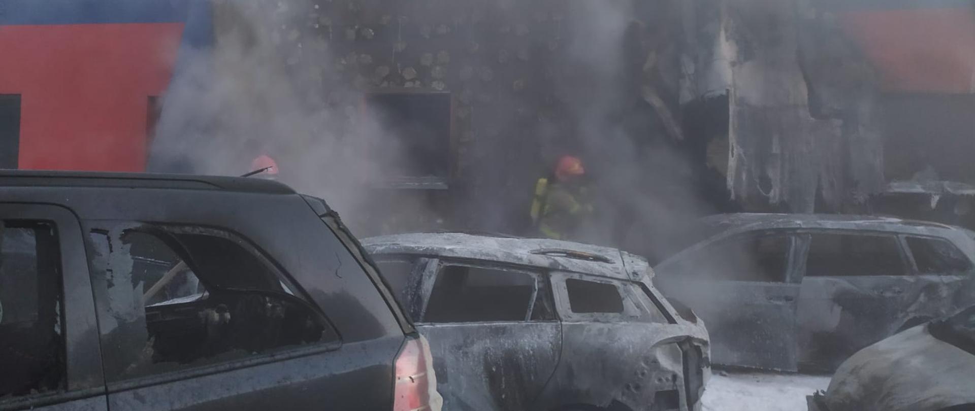 Na zdjęciu widać spalone pojazdy zaparkowane obok budynku warsztatu naprawczego. Na drugim planie uszkodzona zewnętrzna ściana budynku. Poniżej strażacy dogaszający zarzewia ognia.