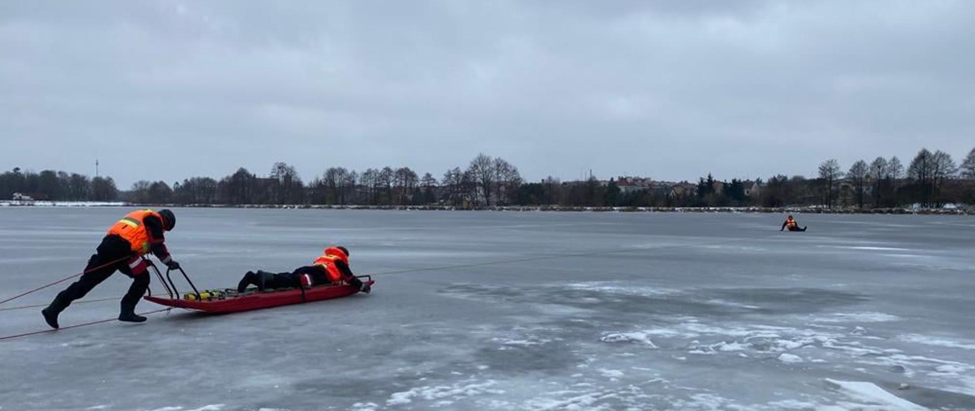 Ćwiczenia na terenie zamarzniętego zalewu miejskiego. Jeden strażak, pcha sanie lodowe na których leży drugi strażak. Obydwaj są ubrani w suchy skafander i kamizelkę ratunkową oraz zabezpieczeni liną. W oddali widać leżącego strażaka, który jest pozorantem.