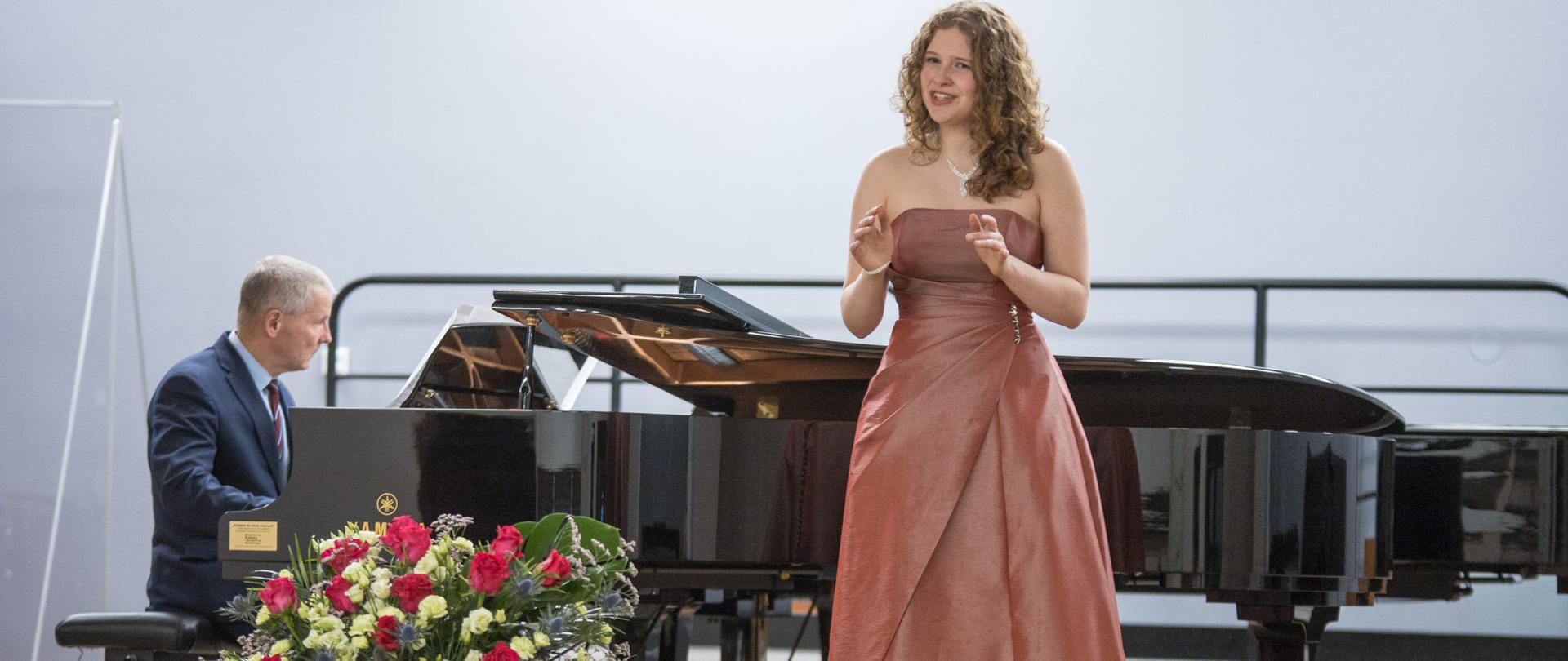 Zdjęcie przedstawia śpiewaczkę Oliwię Ślipek oraz akompaniującego jej na fortepianie Zbigniewa Szewczyka podczas koncertu w auli PSM w Nowym Sączu.